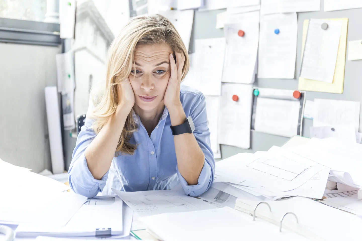 A stressed out woman sits at her desk with her hands on her head and looking at paperwork.