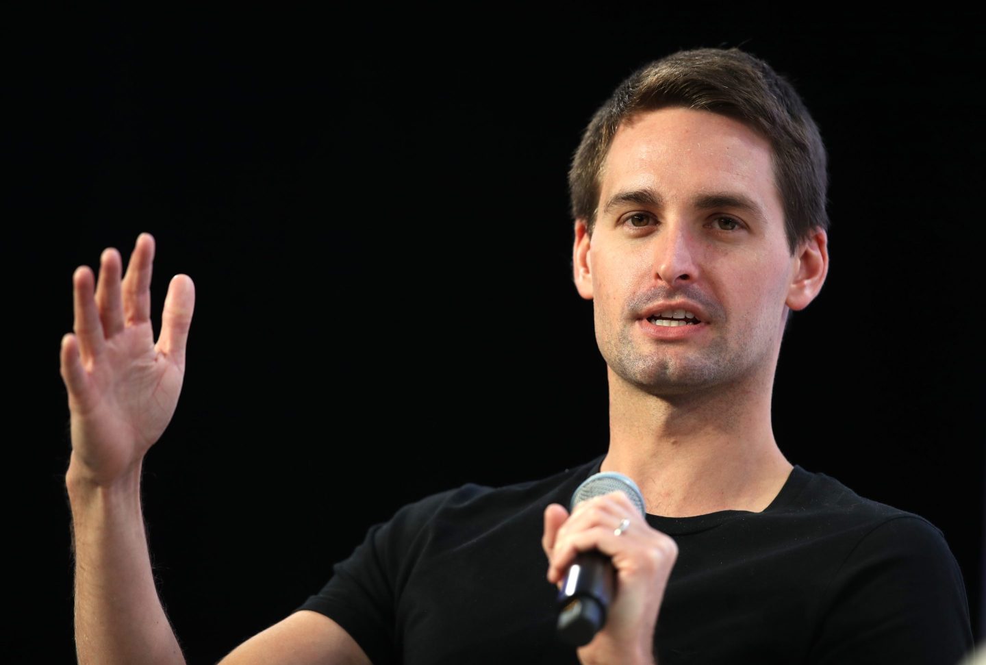 Evan Spiegel wears a black shirt, holds a mic near his face, and holds his hand up