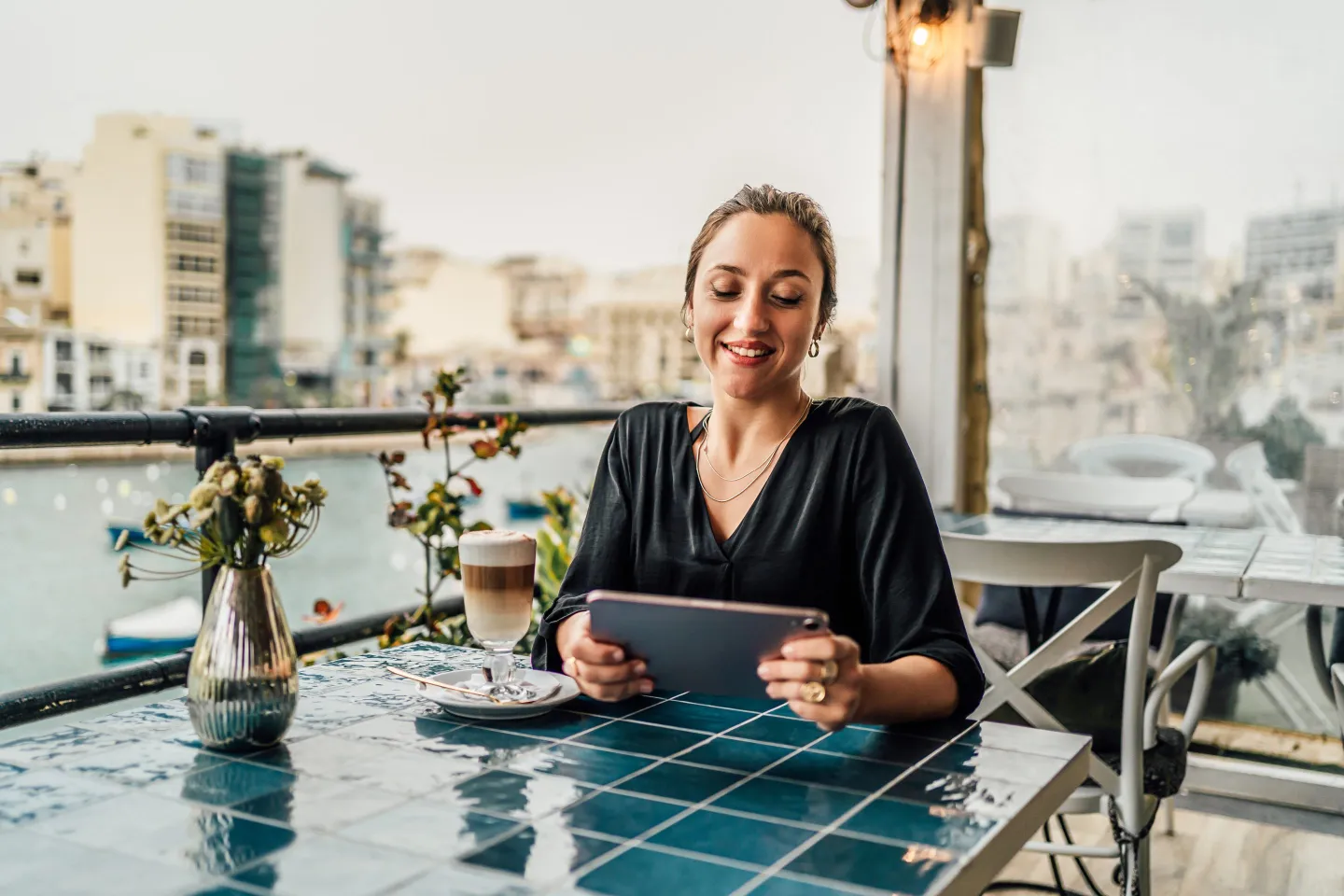 Young cheerful woman using a tablet and drinking coffee in a seaside café in St. Julian's, Malta