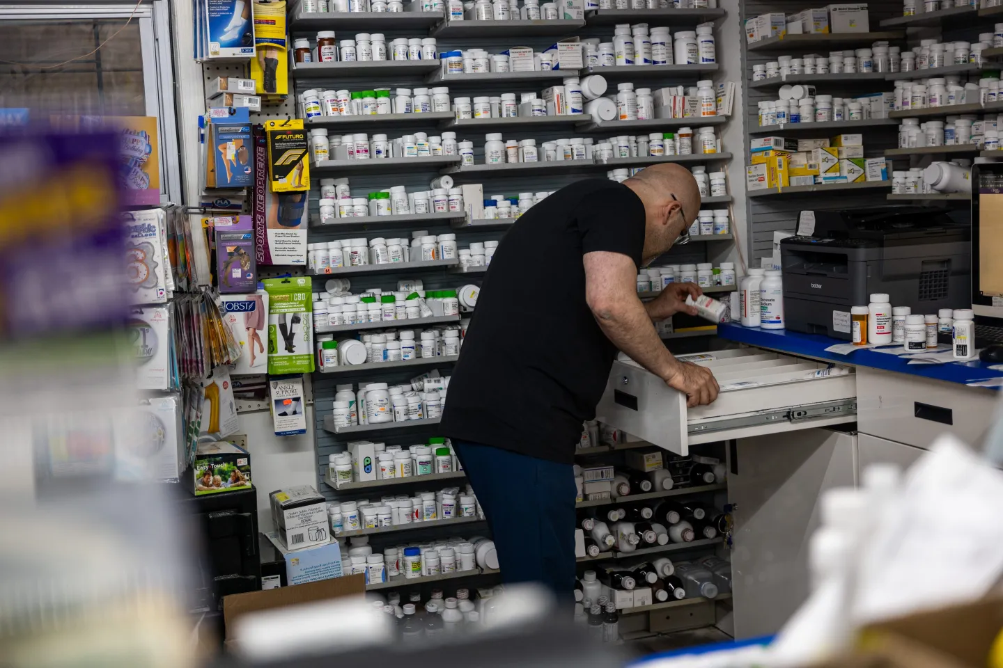 A man leans over and reaches for a bottle while standing in front of a shelf on medications.
