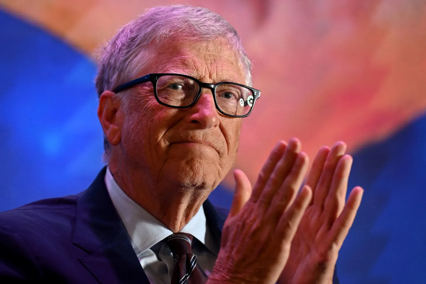 Bill Gates wears a suite and glasses, holding his hands together